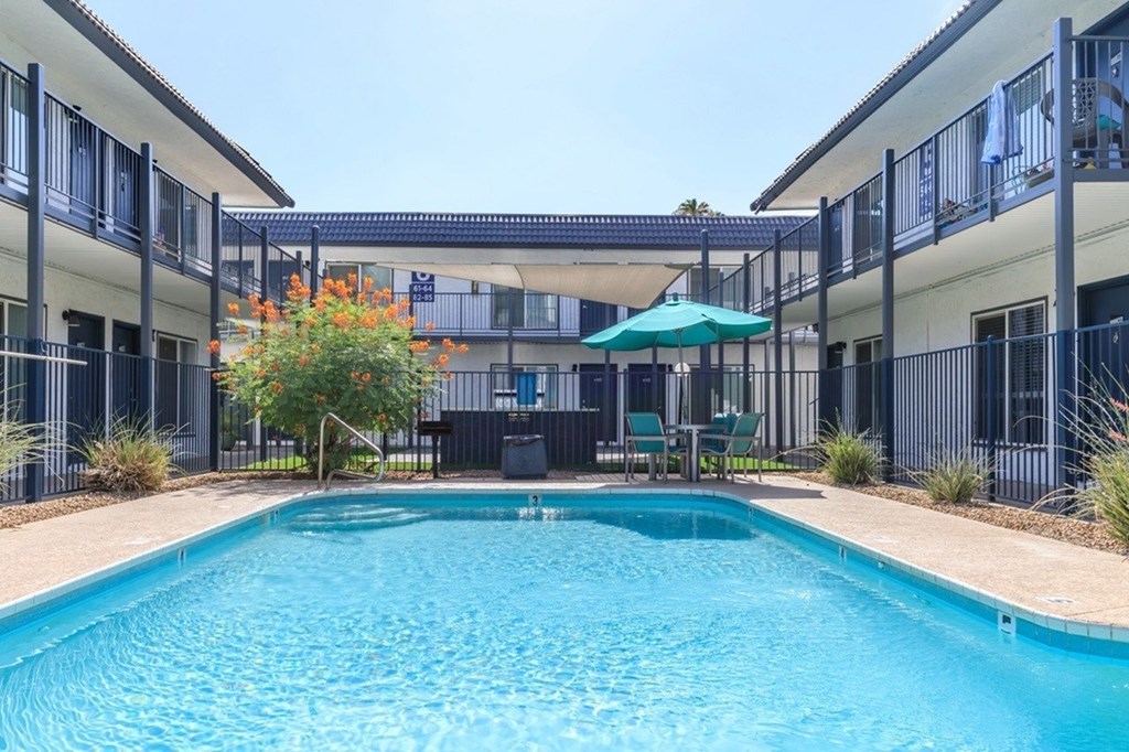 A swimming pool is surrounded by a fence and a patio with chairs and an umbrella.