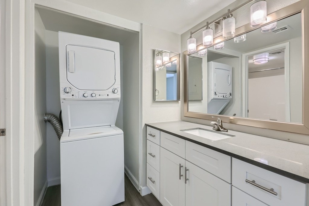 A white washing machine is in a small bathroom with a mirror and lights above it.