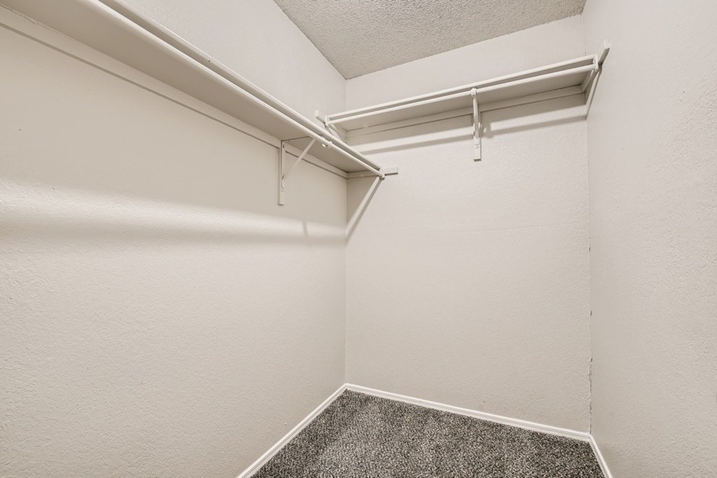 A white wall with a grey carpeted floor and a white ceiling with a white pipe running across it.