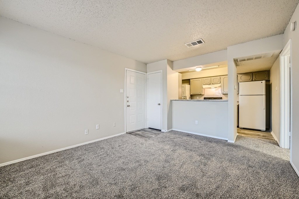 A room with a carpeted floor and a kitchen area in the background.