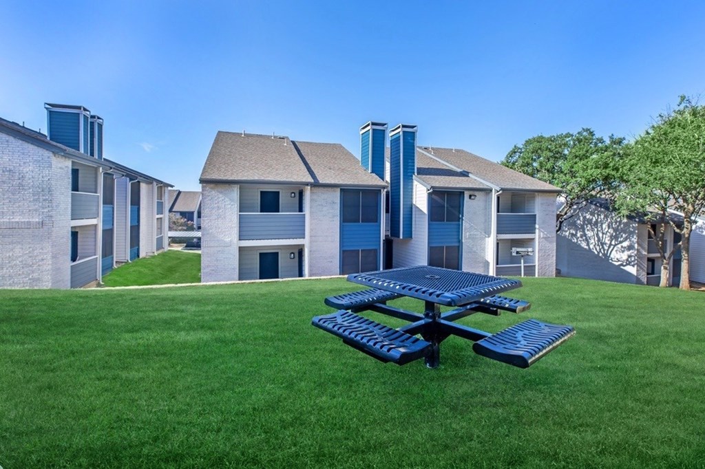 A blue picnic table is in the foreground of a grassy area with apartment buildings in the background.