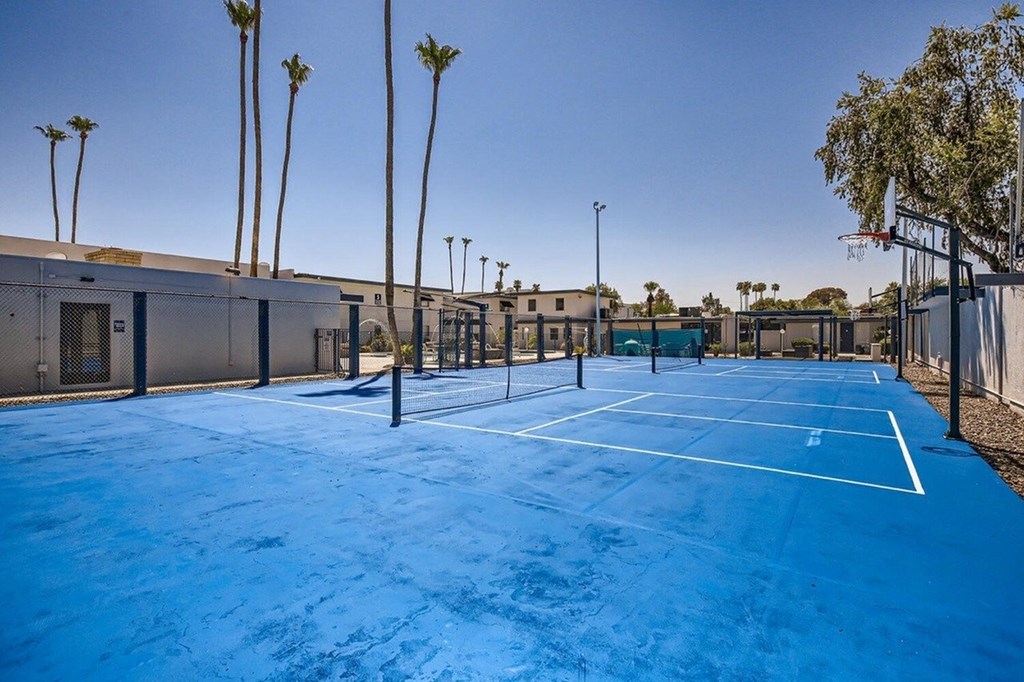 A tennis court with a blue surface and white lines, surrounded by palm trees and buildings.