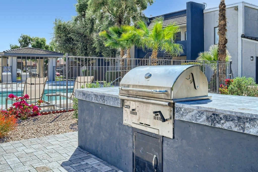 A BBQ grill is in the foreground of a patio area with a pool and palm trees in the background.