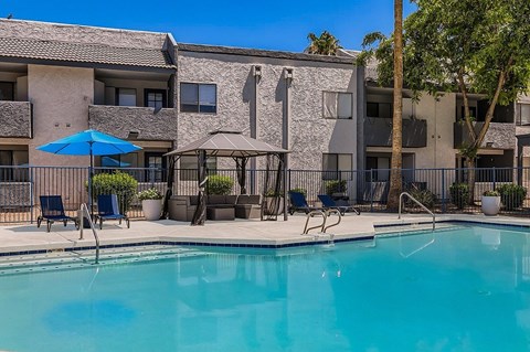 A swimming pool in front of a building with a blue umbrella and chairs.