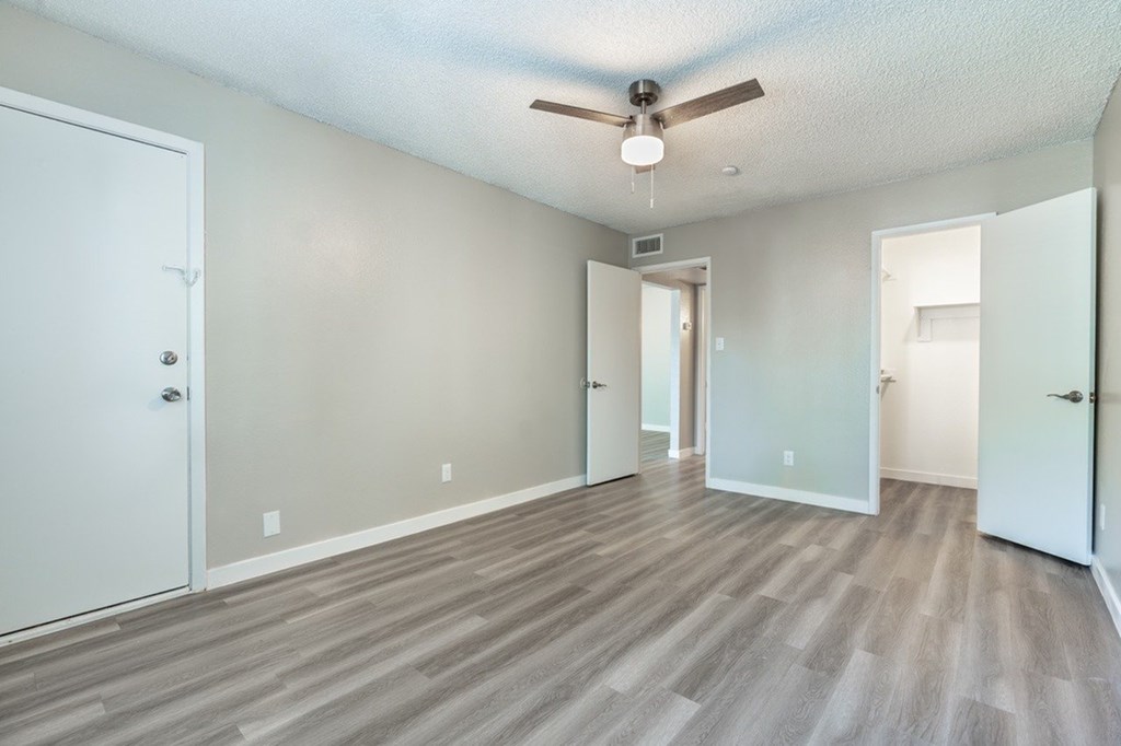 A room with a ceiling fan and light, hardwood floors, and white walls with a door on the left.