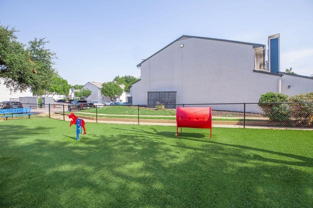 A red object is in the foreground of a grassy area with a building in the background.