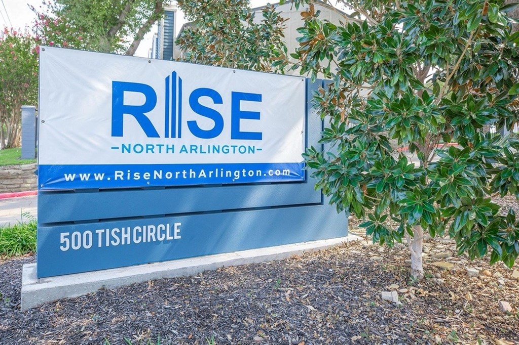 A blue and white sign for RISE North Arlington is displayed in front of a tree.