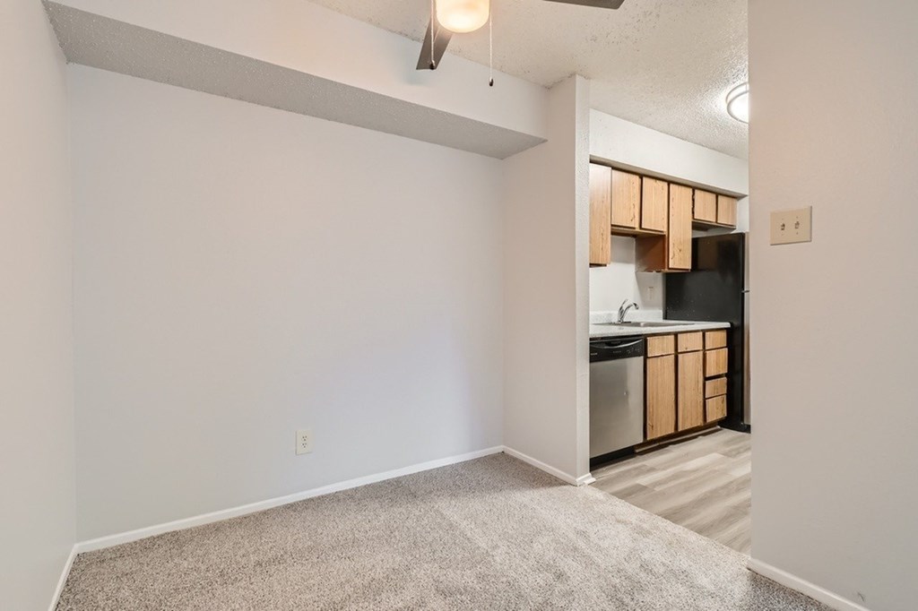 A room with a carpeted floor and a kitchen area in the background.