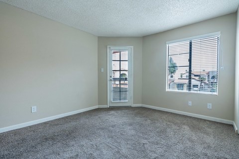 A room with a carpeted floor, a window with blinds, and a door leading to another room.
