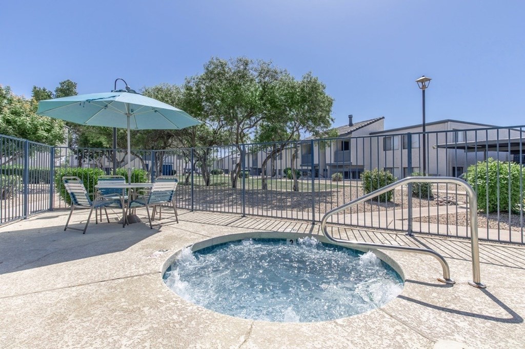 A hot tub sits in the middle of a patio with a blue umbrella and chairs around it.