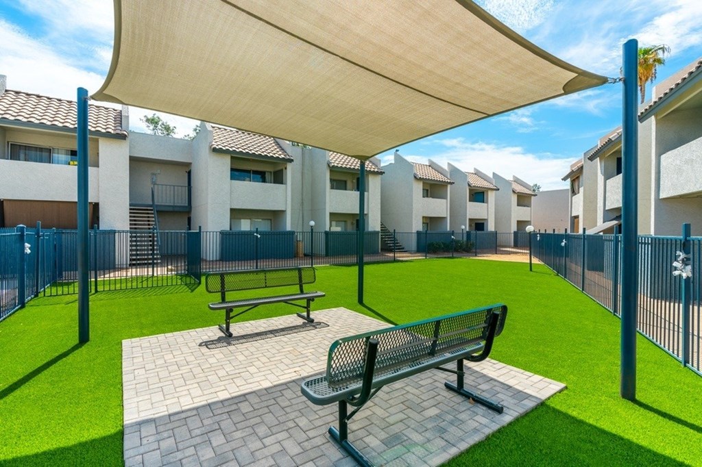 A park area with benches and a canopy is surrounded by a fence and buildings.