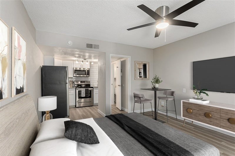A modern bedroom with a large bed, a ceiling fan, and a kitchen area in the background.