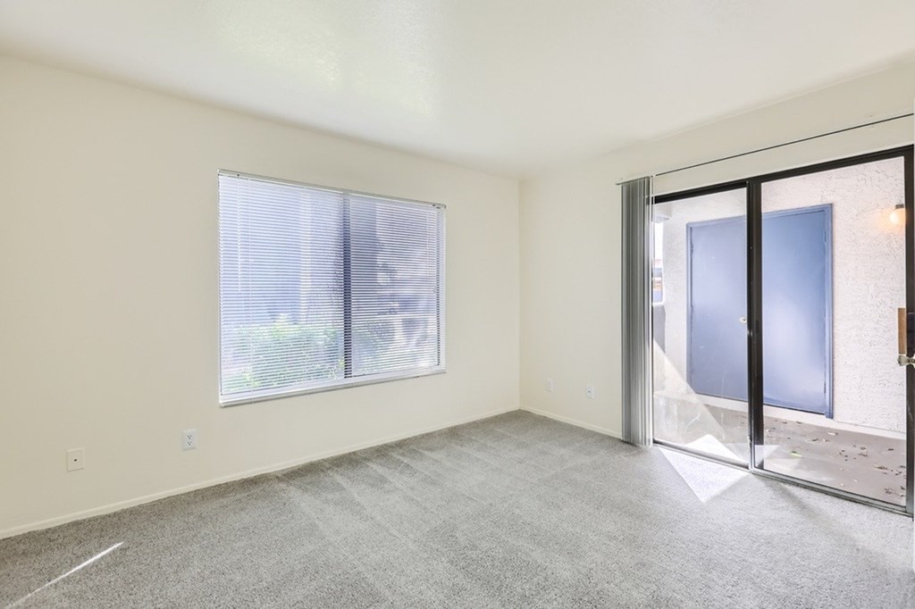 A room with a carpeted floor, a window with blinds, and a sliding glass door leading to a balcony.