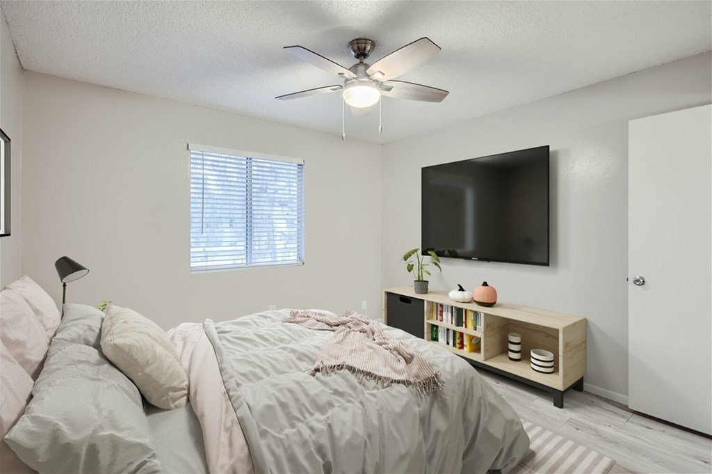 A bedroom with a bed, a ceiling fan, a window, a TV, and a shelf with books and decorative items.