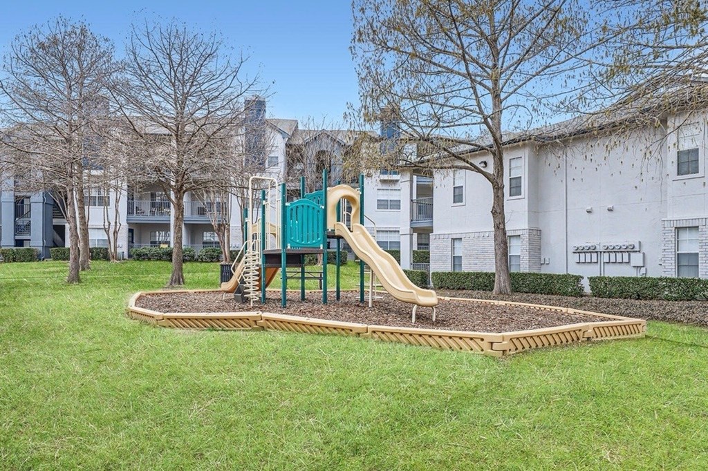 A playground with a green slide in the middle of a grassy area.