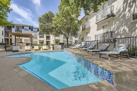 A swimming pool surrounded by lounge chairs and trees in front of apartment buildings.