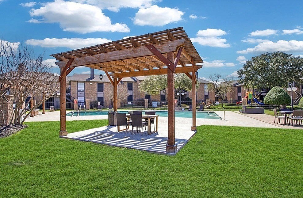 A wooden pergola is over a table in a grassy area.