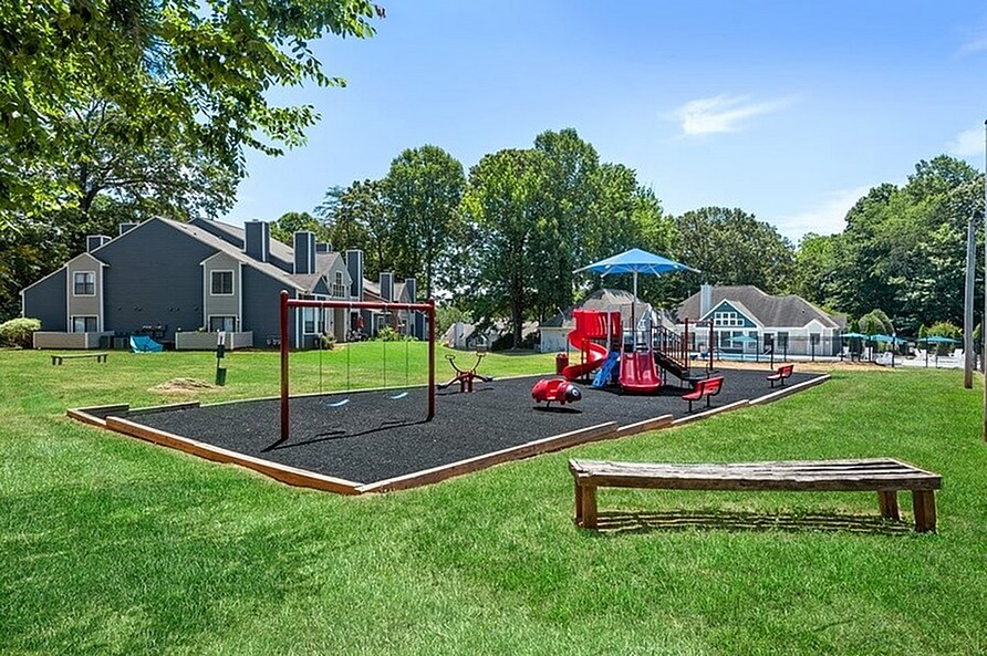 A playground with a slide, swings, and a bench is surrounded by houses and trees.