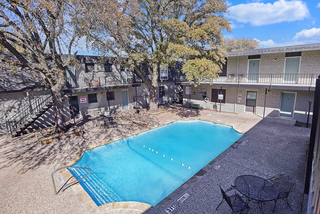 A pool surrounded by trees and a patio with chairs.