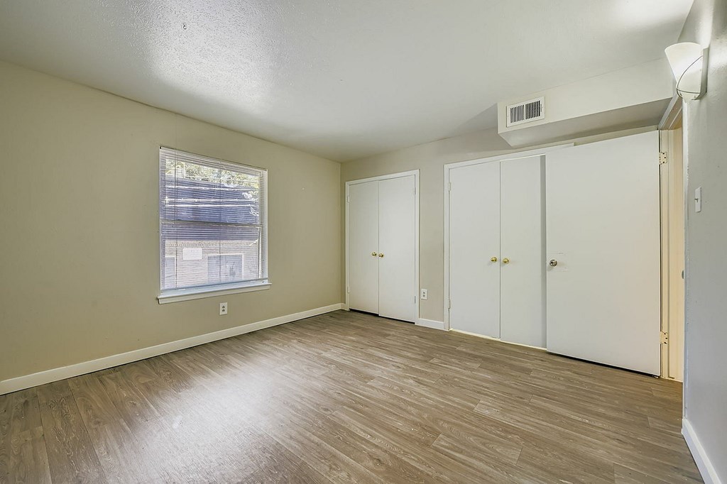A room with a window, a wall-mounted air conditioner, and a wooden floor.