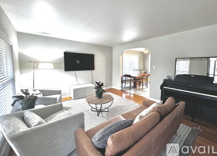 A living room with a brown couch, a coffee table, a television, a piano, and a dining area in the background.