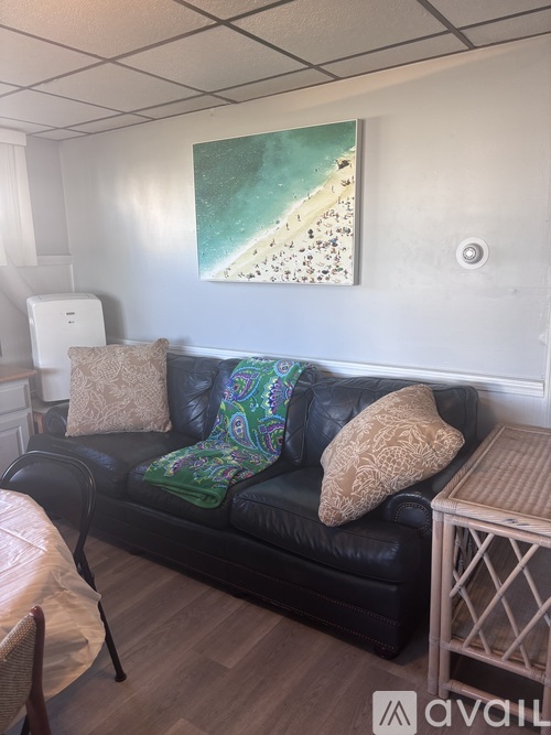A black leather couch with a green and blue throw pillow sits in a room with a painting of a beach scene on the wall.