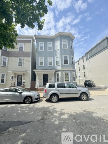 A silver car is parked in front of a grey building with a white car parked in front of a grey building.