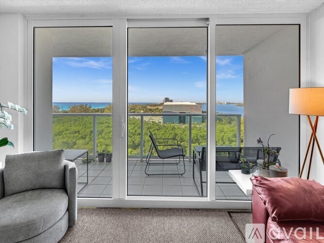 A living room with a grey sofa, a grey armchair, a glass table and a glass door leading to a balcony with a view of the ocean.