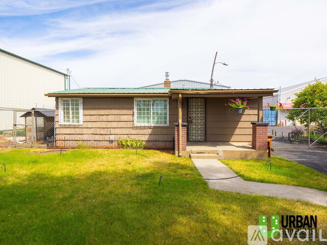 A house with a green roof and a brown exterior is surrounded by a grassy area and a sidewalk.