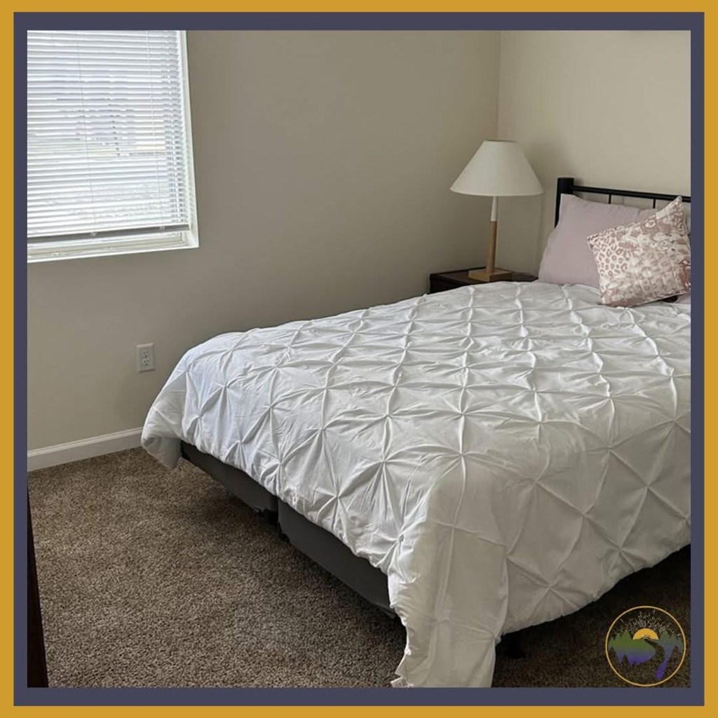 A bed with a white comforter and a pink pillow is in the corner of a room with a lamp and a window.