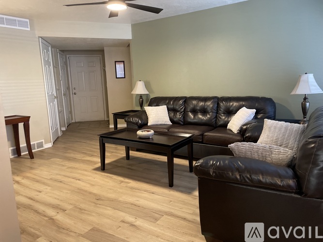 A bed with a white and grey patterned comforter and a purple pillow is in the foreground of a room with wooden flooring and a dark wall.