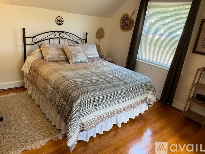 A bed with a striped comforter and pillows sits in a room with wooden floors and a window with curtains.
