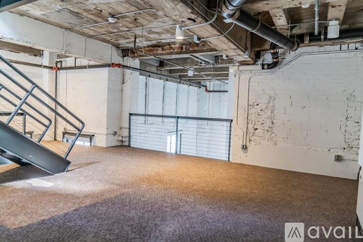 A large, empty room with a carpeted floor, a metal staircase, and exposed pipes on the ceiling.
