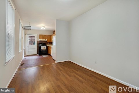 A room with wooden floors and white walls, with a kitchen visible in the background.