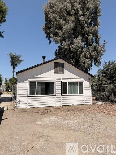 A small house with a white siding and a brown roof is surrounded by trees and has a chain link fence in front of it.