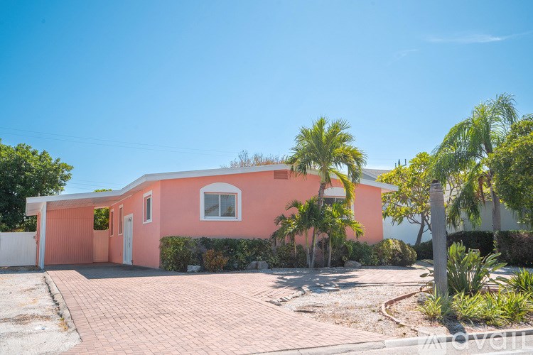 A pink house with a white door and windows is surrounded by greenery and a clear blue sky.