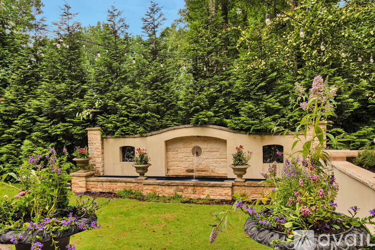A stone building with a balcony is surrounded by greenery and flowers.