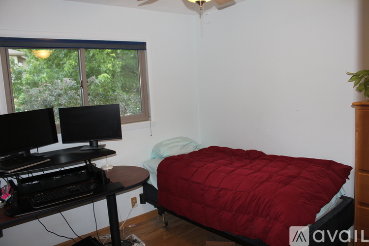 A room with a bed, a desk with a keyboard and two monitors, and a window with a view of trees.