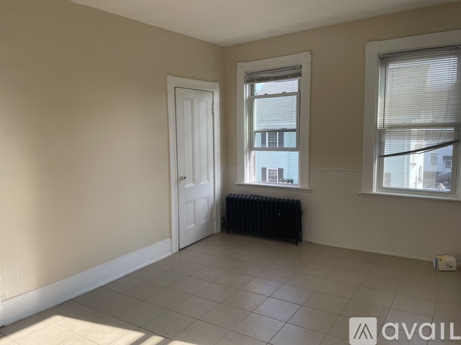 A room with a white door, a window with blinds, and a radiator on the floor.