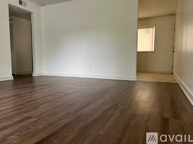 A room with wooden flooring and white walls, with a door on the left and a window on the right.