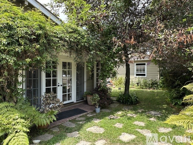A small white house with a porch surrounded by green plants.