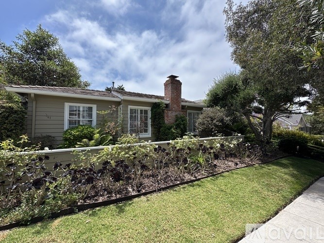 A house with a grey exterior and a red brick chimney is surrounded by a well-kept lawn and a hedge.