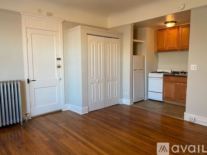 A room with wooden floors and white walls, with a kitchen area visible in the background.