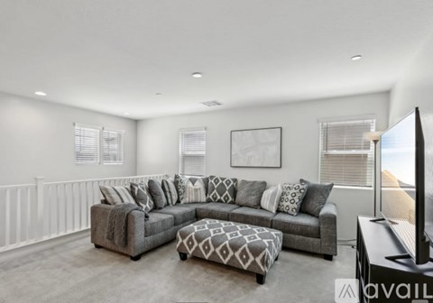 A living room with a grey sofa and ottoman, a flat screen TV, and a large window with blinds.