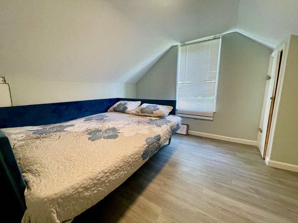 A bed with a blue headboard and a white comforter with blue floral patterns sits in a room with light wood flooring and a window with blinds.