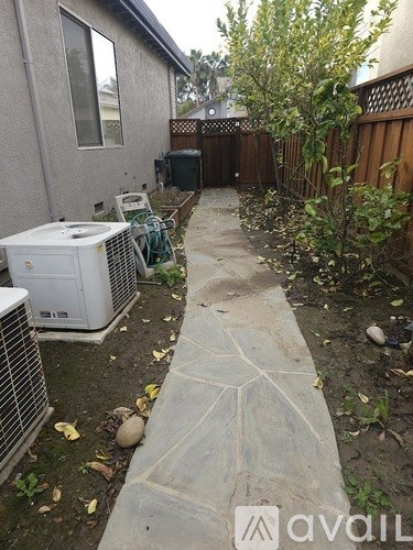 A concrete pathway leads from a house to a yard with a white air conditioning unit and a green lawn chair.