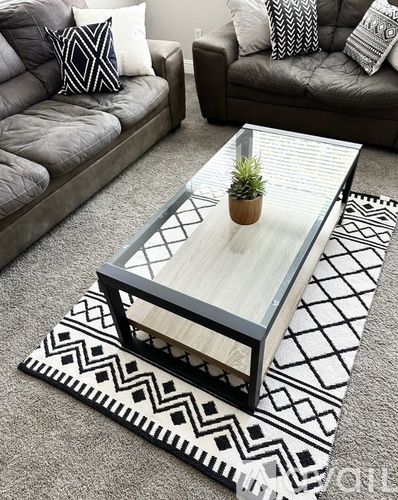 A glass coffee table with a wooden top and black frame sits on a patterned rug in a living room.