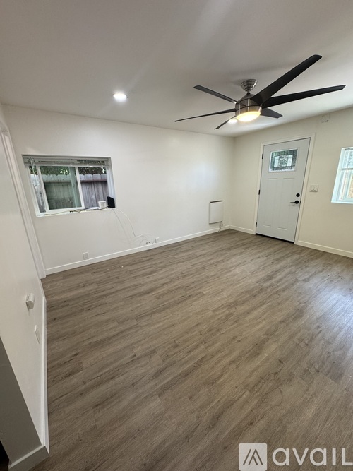 A room with a ceiling fan and light, wooden flooring, and a window with blinds.