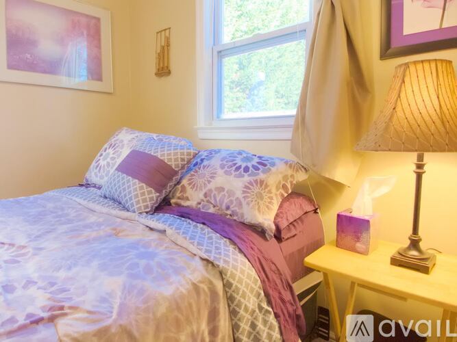 A bed with a purple and white comforter and matching pillows sits next to a lamp and a window.