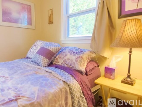 A bed with a purple and white comforter and matching pillows sits next to a lamp and a window.